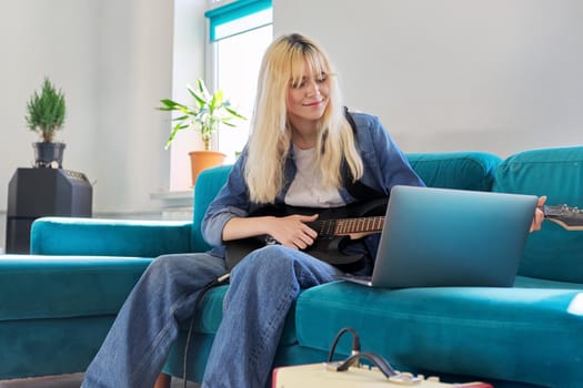 Fashionable female teenager playing electric guitar at home. Girl hipster blonde sitting with an electric guitar on sofa in living room. Music, creativity, adolescence, hobbies and leisure concept