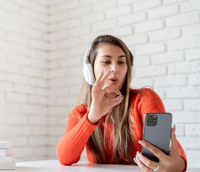 Social media. Young caucasian woman wearing earphones chatting on mobile showing okay sign