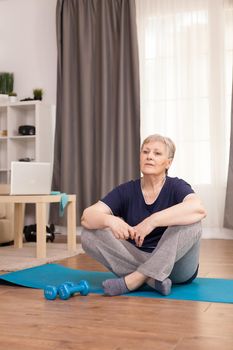 Woman sitting on yoga mat waiting for the wellness trainer. Old person pensioner online internet exercise training at home sport activity with dumbbell, resistance band, swiss ball at elderly retirement age