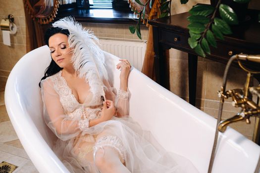 the bride, dressed in a boudoir transparent dress and underwear, lies in a vintage bathroom with a white feather in her hands, The morning of the bride.