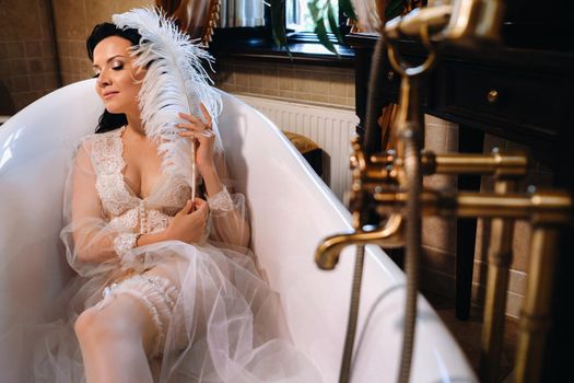 the bride, dressed in a boudoir transparent dress and underwear, lies in a vintage bathroom with a white feather in her hands, The morning of the bride.