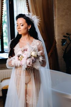 the bride, dressed in a boudoir transparent dress and underwear, sits near a vintage bath with a feather in her hands, The morning of the bride.