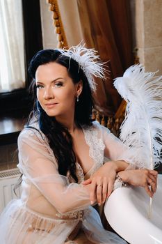 the bride, dressed in a boudoir transparent dress and underwear, sits near a vintage bath with a feather in her hands, The morning of the bride.