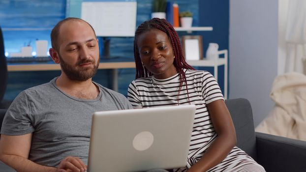 Young interracial couple waving at video call camera on laptop while talking to friends in living room. Married mixed race people using online remote communication on conference