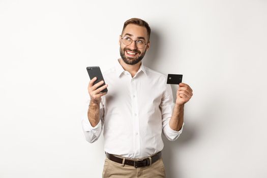 Business and online payment. Image of handsome man thinking while holding credit card and smartphone, standing against white background.