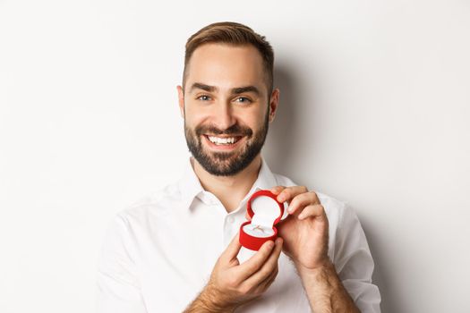 Close-up of happy handsome man making a proposal, holding wedding ring in box and smiling, asking to marry him, white background.