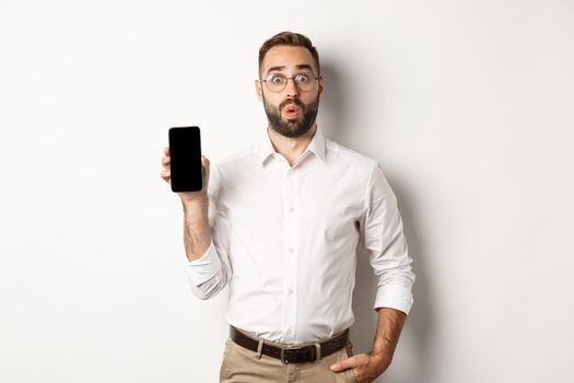 Surprised handsome manager in glasses, looking curious and showing mobile screen, standing over white background.