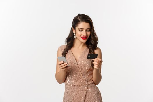 Online shopping and christmas concept. Elegant young woman paying for internet purchase with smartphone and credit card, standing in luxury dress over white background.