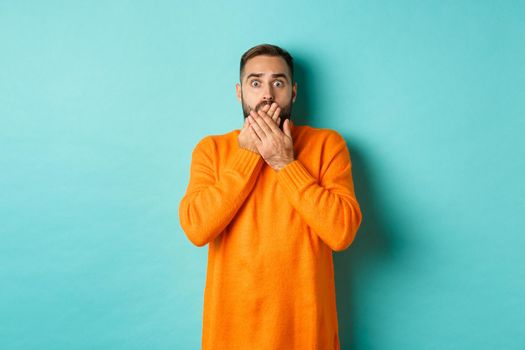 Startled man looking at horrible scene, gasping and stare scared, standing in orange winter sweater, studio background.