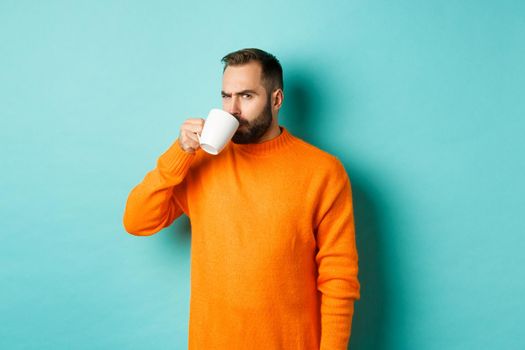 Image of guy drinking coffee and looking suspicious at camera, stare at something strange, standing in orange sweater over turquoise background.