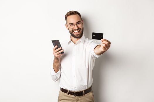 Business and online payment. Excited man showing his credit card while holding smartphone, standing satisfied against white background.