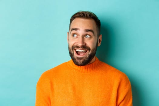 Close-up of handsome caucasian man smiling, looking left with surprised face, staring at logo, wearing orange sweater, standing against turquoise background.