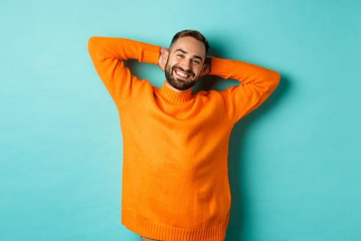Handsome man relaxing, resting and smiling, holding hands behind head with carefree face, standing against light blue background.