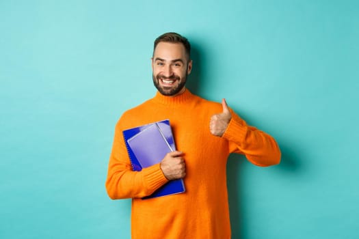 Education. Smiling bearded man holding notebooks and smiling, going on courses, showing thumbs-up in approval, like something, standing over turquoise background.