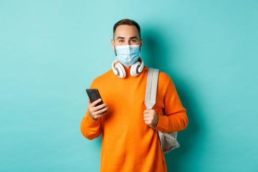 Young man in face mask using mobile phone, holding backpack, standing against light blue background. Copy space