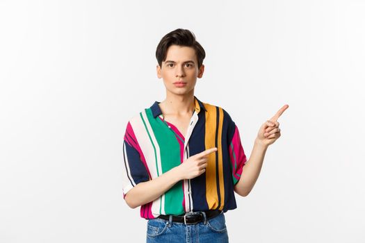 Portrait of young handsome man pointing fingers at upper right corner, showing logo, standing over white background.