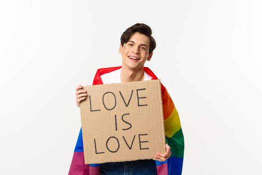 Smiling gay man activist holding sign love is love for lgbt pride parade, wearing Rainbow flag, standing over white background.