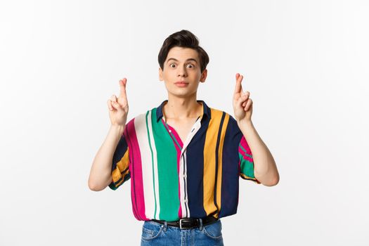 Hopeful young androgynous man cross fingers for good luck, making wish, waiting for something, standing over white background.