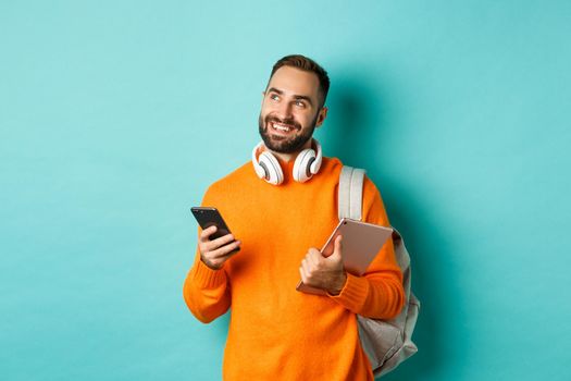 Handsome man student with headphones and backpack, holding digital tablet and smartphone, looking dreamy at upper left corner, standing against turquoise background.