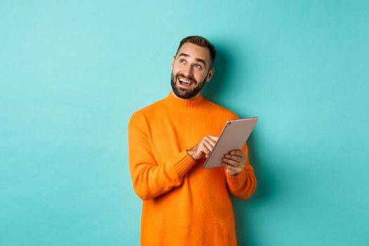 Handsome man thinking and using digital tablet, shopping online and pondering ideas, looking at upper left corner, standing over light blue background.