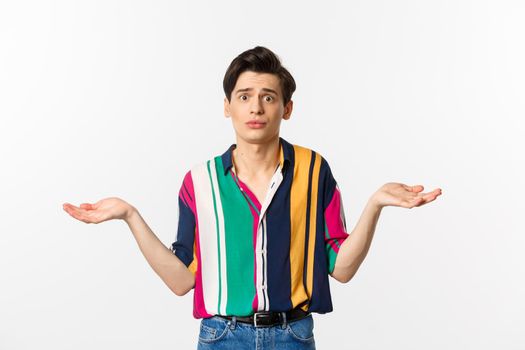 Sad handsome guy shrugging shoulders and looking confused, dont know what to do, holding something in hands, standing over white background.