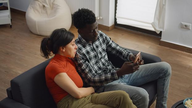 Married interracial couple taking selfie with smartphone while sitting together on sofa at home. Multi ethnic man and woman using modern device for joyful picture in living room
