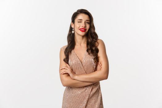 Beautiful woman in evening dress and red lipstick, winking at camera and smiling, celebrating New Year party, standing over white background.