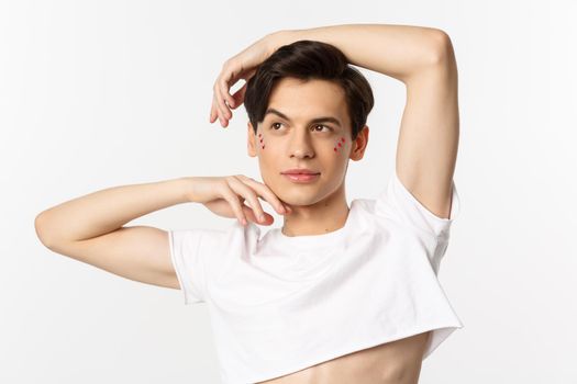 Close-up of beautiful androgynous man in crop top posing for camera, standing against white background.