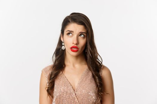 Concept of New Year celebration and winter holidays. Close-up of elegant woman with red lips and dress, looking at upper left corner and thinking, standing over white background.