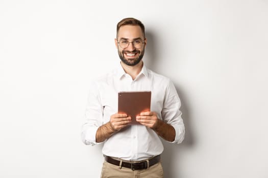 Handsome bearded man in glasses using digital tablet, smiling satisfied, standing against white background.