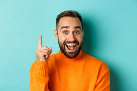 Close-up of handsome man having an idea, raising finger up and smiling excited, standing in orange sweater.