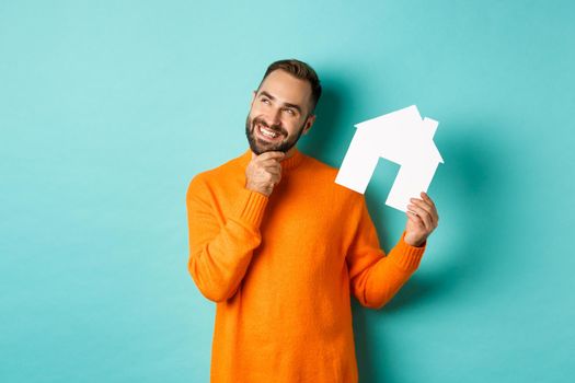 Real estate concept. Handsome adult man in orange sweater thinking about buying flat or renting, holding house maket and looking thoughtful, standing over blue background.