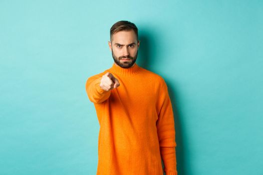Confident man blame you, frowning and pointing finger at camera, accuse person, judgemental stare, standing over light blue background.