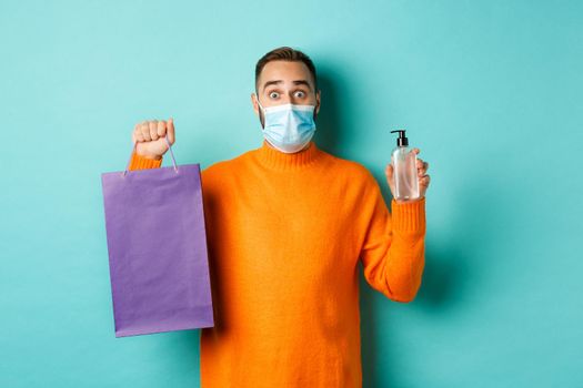 Coronavirus, pandemic and lifestyle concept. Man in face mask showing shopping bag and hand sanitizer, standing over turquoise background.