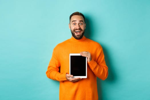 Technology. Excited adult man in orange sweater showing digital tablet screen, staring amazed at camera, standing over turquoise background.