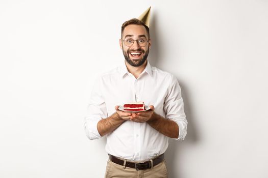 Holidays and celebration. Happy man having birthday party, making wish on b-day cake and smiling, standing against white background.