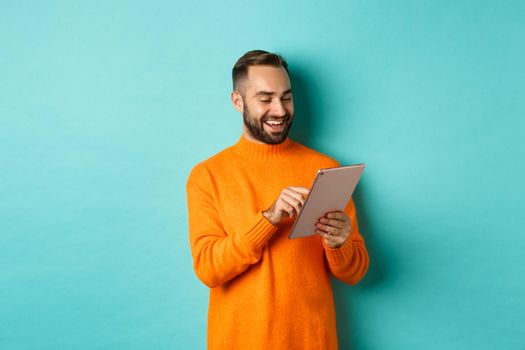 Handsome adult man working on digital tablet and smiling, shopping online, standing over light blue background.