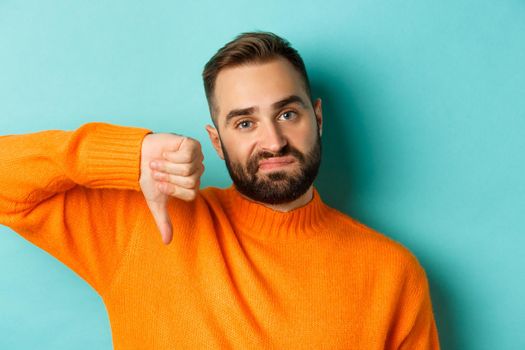 Close-up of disappointed caucasian man with beard, showing thumb down and grimacing displesed, negative feedback, standing over light blue background.