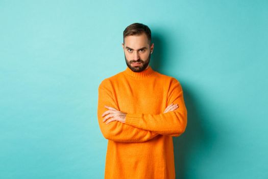 Offended man looking angry at you, cross arms on chest and stare mad, standing in orange sweater against turquoise background.