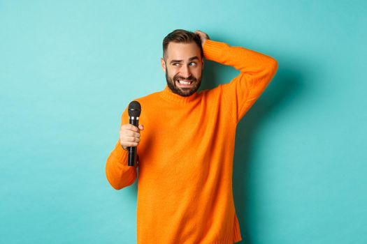 Shy and awkward adult man scratching head, holding microphone before singing, standing over blue background.