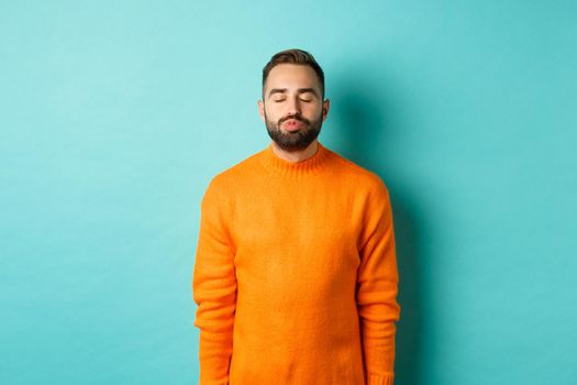 Waist up shot of young man pucker lips and close eyes, waiting for kiss, standing in orange sweater against light blue background.