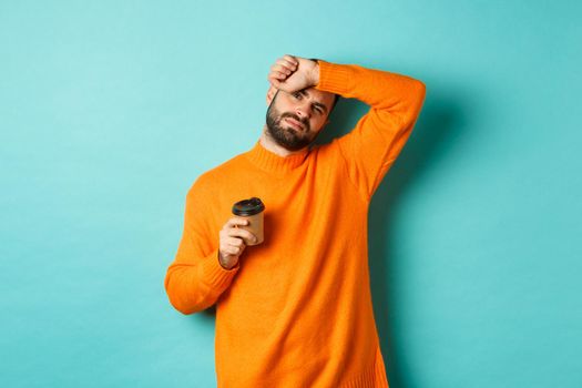 Tired adult man taking break from work and drinking coffee, wipe sweat off forehead and looking exhausted, standing in orange sweater over turquoise background.