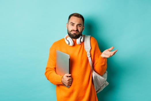 Image of troubled young man with headphones and backpack, shrugging confused and holding laptop, standing over light blue background.