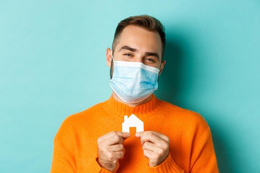 Real estate and coronavirus pandemic concept. Close-up of adult man in medical mask holding small paper house maket and smiling, searching for apartment, light blue background.
