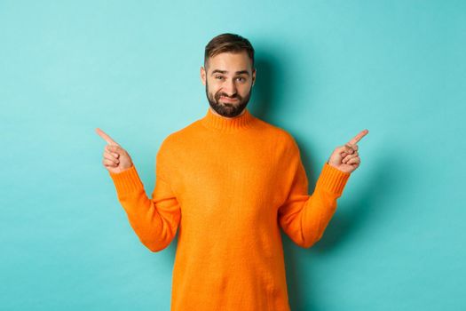 Doubtful and indecisive man pointing fingers sideways, showing different sides, two choices, looking skeptical, standing over light blue background.