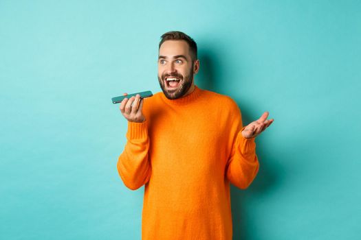 Happy man talking on speakerphone, gesturing and recording voice message on mobile phone, standing in orange sweater over light blue background.