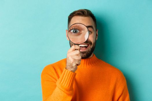 Funny man looking through magnifying glass, searching or investigating something, standing in orange sweater against turquoise background.