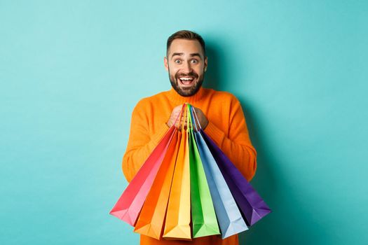 Excited adult man holding shopping bags and smiling, going to mall, standing over turquoise background.