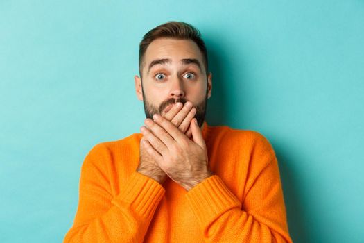 Close-up of shocked caucasian man covering his lips and mouth, staring astounded at camera, standing against turquoise background.