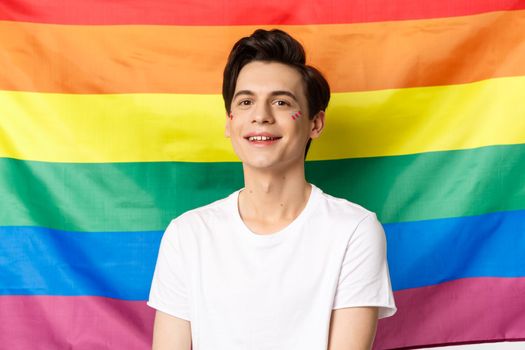 Beautiful gay man with glitter on face, smiling happy and proud at camera, standing against rainbow pride flag, lgbtq community rights and people concept.